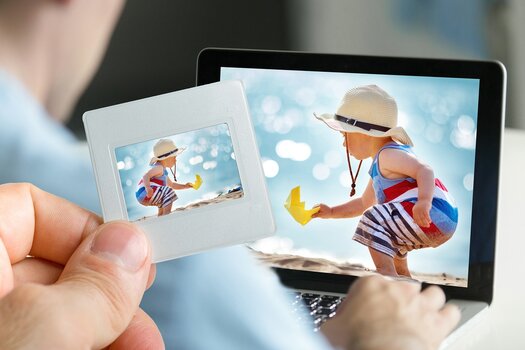Bild eines am Strand spielenden Kleinkinds wird auf Laptopmonitor betrachtet. Im Vordergrund hält eine Hand das Dia mit demselben Motiv in die Kamera.