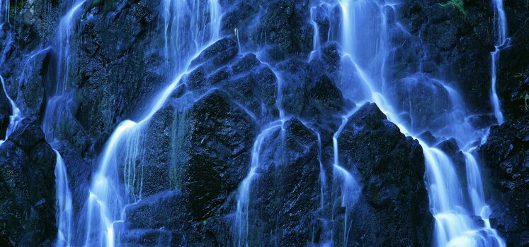 Foto vieler kleiner Wasserfälle nebeneinander, im Hintergrund ist die Felswand gut sichtbar. Vorherrschende Farbatmosphäre in Blautönen