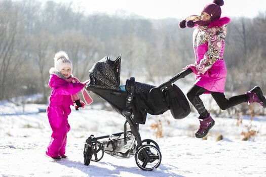 Zwei Kinder mit pinker Winterkleidung vor Schneelandschaft, zwischen ihnen ein Kinderwagen. Das ältere Mädchen rechts wird beim Hochspringen fotografiert., das kleinere Kind links lächelt in die Kamera.