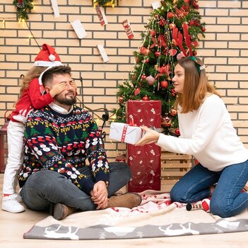 Familie in weihnachtlicher Kleidung sitzt auf dem Boden vor einem kleinen Weihnachtsbaum. Das Kind steht hinter dem Vater und hält ihm die Augen zu, während die Mutter ihm ein eingepacktes Geschenk hinhält. 