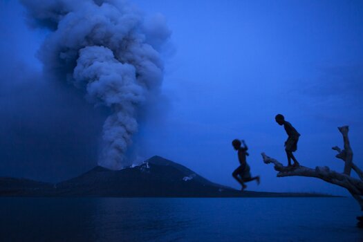 Zwei Kinder springen von einem Baumstamm ins Wasser. Im Hintergrund ein rauchender Vulkan. Das Bild ist in Blautönen gehalten.