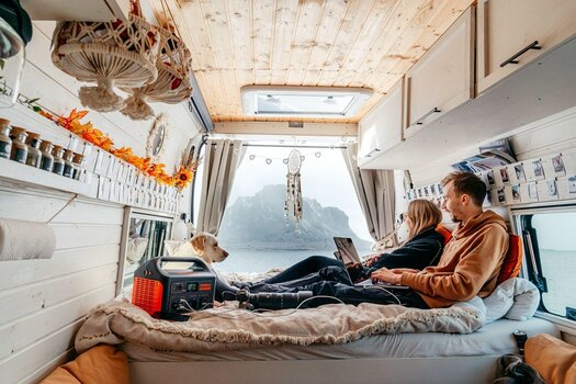 Paar mit Hund sitzt im Wohnwagen oder Wohnmobil auf dem Bett und bedient Laptop und Handy. Die Geräte werden durch einen Jackery Explorer geladen. 