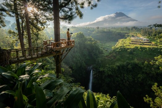 Mann mit Kamera steht auf einer hölzernen Aussichtsplattform mit beeindruckendem Blick über indonesische Landschaft