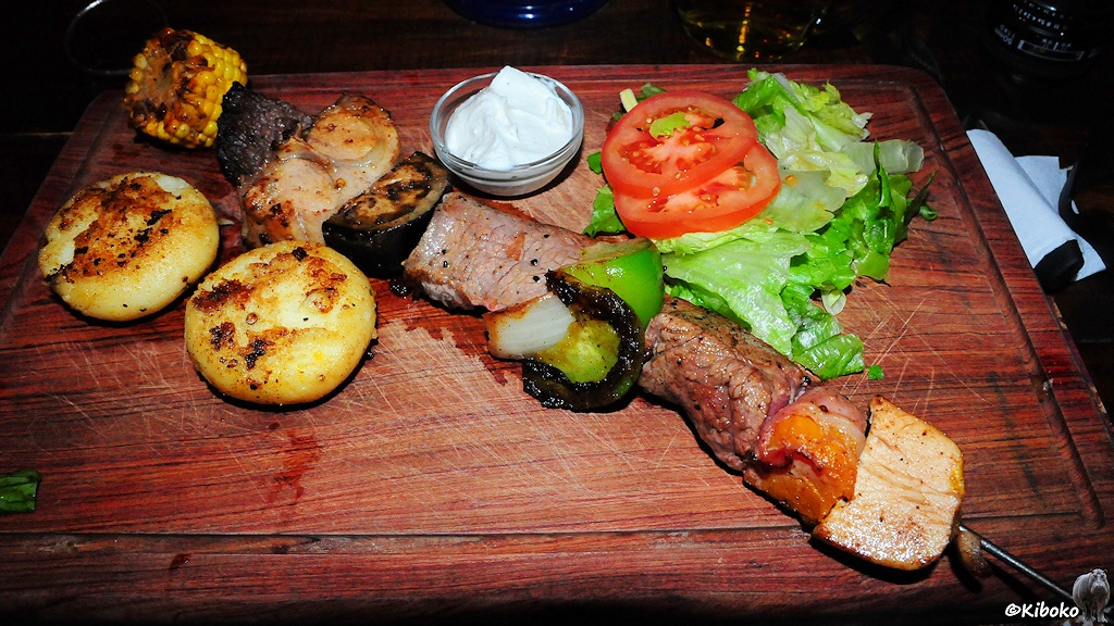 Ein gegrillter Fleischspieß mit vier Sorten Fleisch, ein Salatblatt mit Tomatenscheiben und zwei Kartoffeln liegen auf einem Holzbrett.