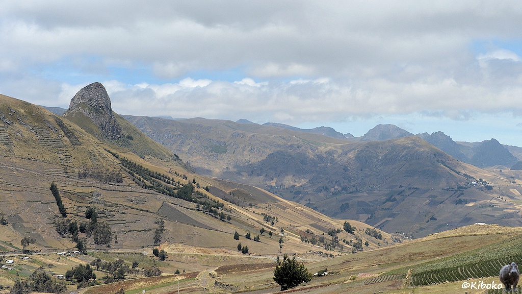 Das Bild zeigt eine Berglandschaft mit einem Tal. Im Tal sind ein paar kleine Häuser. Auf der rechten Hangseite sind Felder. Auf der linken Hangseite ist ein hoher Granitfelsen. Im Hintegrund ist ein größeres Tal mit weiteren hohen Bergen.