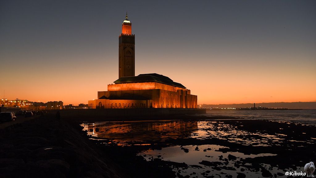 Das Bild zeigt eine Moschee mit rechteckigen Turm am Meer. Am Horizont ist ein oranger Lichtschimmer.