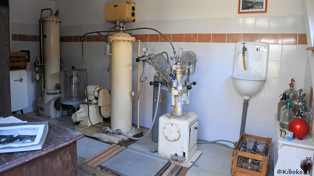 Das Bild zeigt die Abfüllanlage für Sodawasser. Vor einer Wand mit weißen Fliesen stehen zwei beige Tanks mit Pumpen. Rechts ist ein Waschbecken. Auf dem Boden stehe eine Holzkiste mit 9 Glasflaschen.