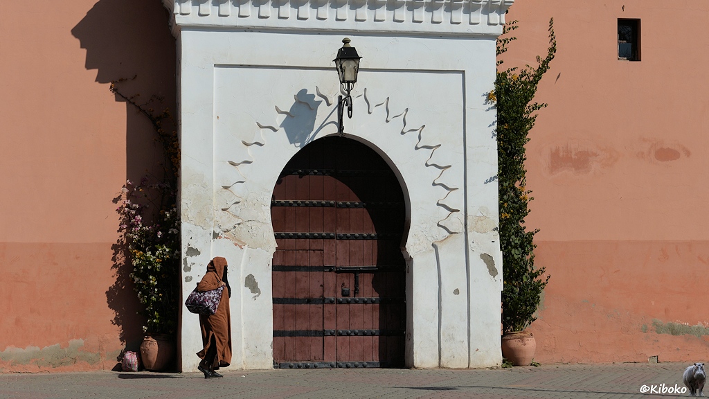 Eine Frau in braunem Kaftan passiert eine große, verzierte Tür an einer hohen rosafarbenen Wand