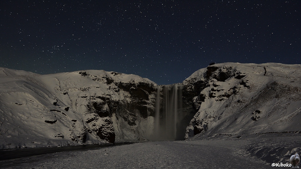 Das Bild zeigt einen 60m hohen Wasserfall, der in mehreren parallelen Strömen herunterfällt, in einer nächtlichen Schneelandschaft. Um den Wasserfall sind schwarze Felsen. Am linken Bildrand verschwindet das abfließende Wasser als kleiner Fluss. Im Vordergrund ist eine ebene festgetrampelte Schneefläche. Darüber leuchten die Sterne an einen dunkelblauen Nachthimmel.