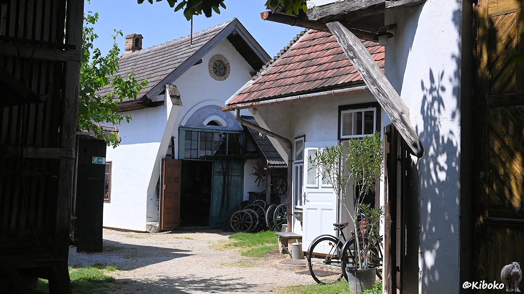 Das Bild zeigt einen Kiesweg zwischen mehreren kleinen einstöckigen Gebäuden. Vor den Gebäuden stehen alte Fahrräder. Vor dem zentralen Gebäude stehen hölzerne Wagenräder.