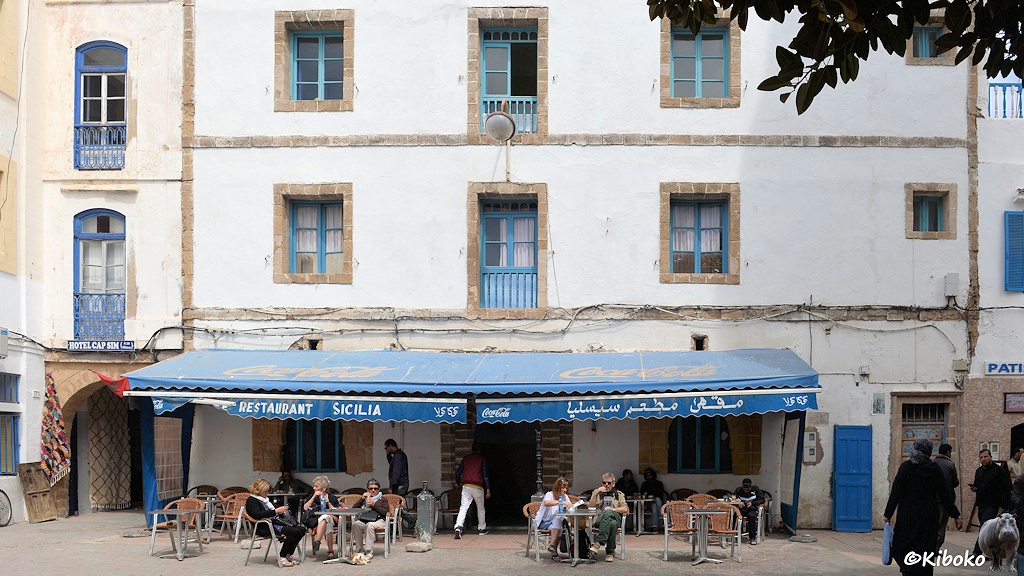 Das Bild zeigt ein dreigeschossiges weißes Haus mit blauen Fenstern und Türen, die mit hellbraunem Sandstein eingefasst sind. Das Gebäude ist ein Restaurant. Unter einer blauen Makise sitzen Touristen in braunen Korbsesseln vor dem Gebäude.