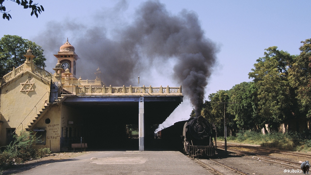 Eine Dampflok fährt mit viel Qualm am Bahnsteig eines beigefarbenes Bahnhofsgebäudes vorbei
