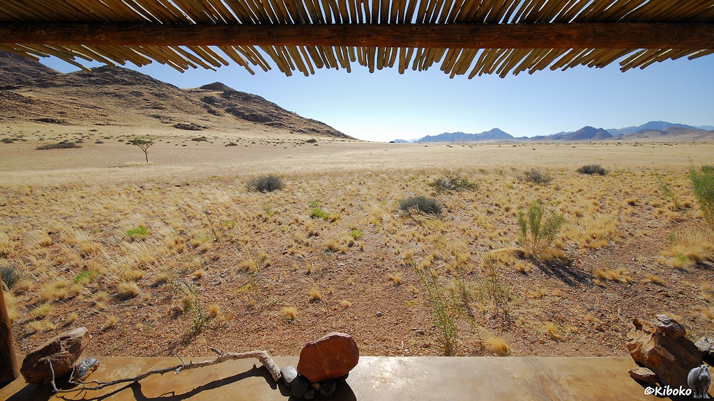Blick von der Veranda auf eine trockene Grasfläche mit Hügeln und Bergen im Hintergrund. Oben ragt der Sonnenschutz aus Bambussstangen ins Bild.