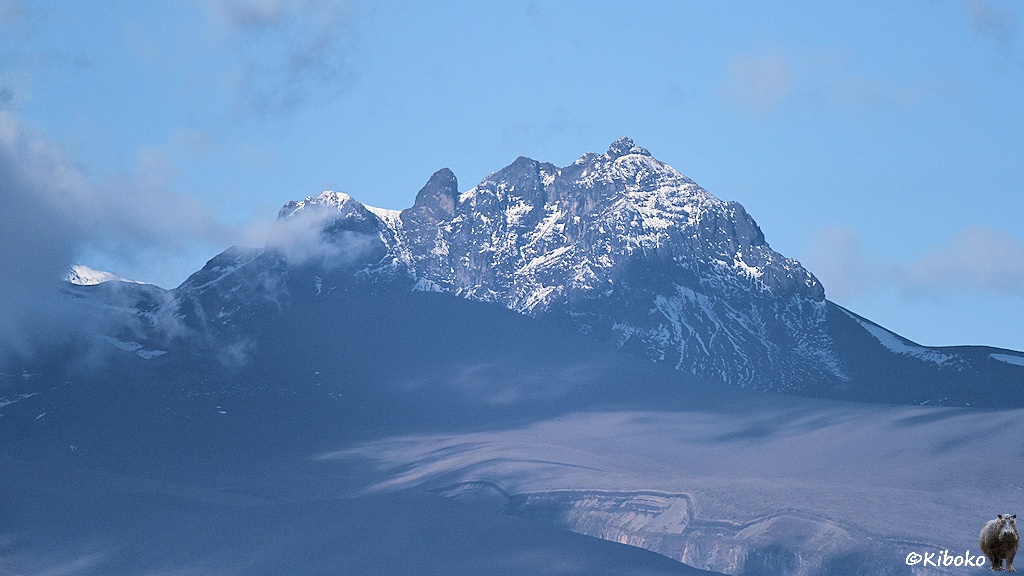 Das Bild zeigt ein felsiges Bergmassiv mit Schnee in den Schluchten. Darunter ist ein baumlose Schotterwüste, die teilweise im Schatten einer Wolke liegt. Am linken Bildrand ist der Anstieg zu einem höheren Berg, der in den Wolken liegt.