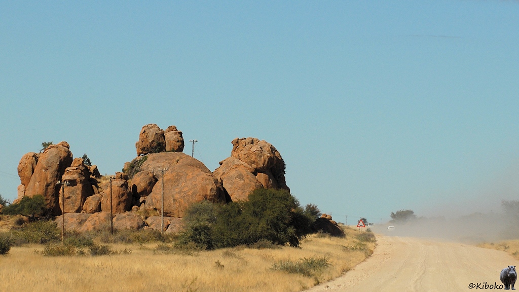 Eine Schotterpiste führt links an einer Felsformation aus rundlichen brauenen Felsen vorbei.