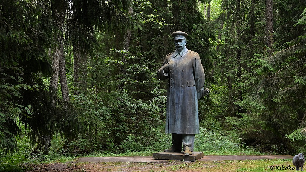 Das Bild zeigt eine überlebensgroße Skulptur von Stalin im langen Mantel und Schirmmütze. Die rechte Hand hat er wie Napoleon in den Mantel gesteckt. Das Kupferoxid und rostfarbene Denkmal steht mitten im Wald zwischen hohen Nadelbäumen. .