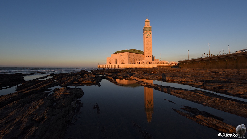 Das Bild zeigt eine Moschee mit viereckigen turm im Abendlicht am Meer. Bei Ebbe ragen Felsen zwischen kleinen Wasserflächen heraus. Darin spiegelt sich der Turm der Moschee. Am rechten Rand ist eine hohe Schutzmauer gegen das Meer.