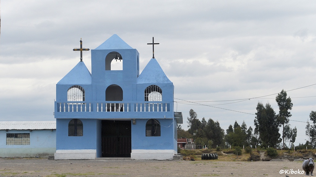 Das Bild zeigt ein kleines Blaues Kirchlein. Neben der braunen Eingangstür ist auf beiden Seiten ein kleines Bogenfenster. Darüber ist Balkon mit weißem Geländer. Darüber sind zwei kleine Räume mit pyramidenförmigem Dach. Der Glockenturm mit zwei Glöckchen ist eine etwag höher und ebenfalls durch ein pyramidenförmiges Dach abgeschlossen. Links ist ein hellblauer Anbau.