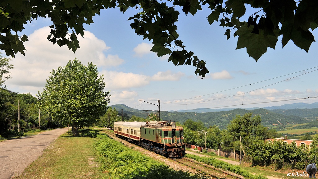 Das Bild zeigt eine alte, grüne, kastenförmige Elektrolokomotive mit zwei kurzen weißen Personenwagen. Parallel zur Bahn fürht eine Straße mit schattenspenden Bäumen. Blätter ragen oben in das Bild hinein. Im Hintergrund sind bewaldete Berge des Kaukasus.