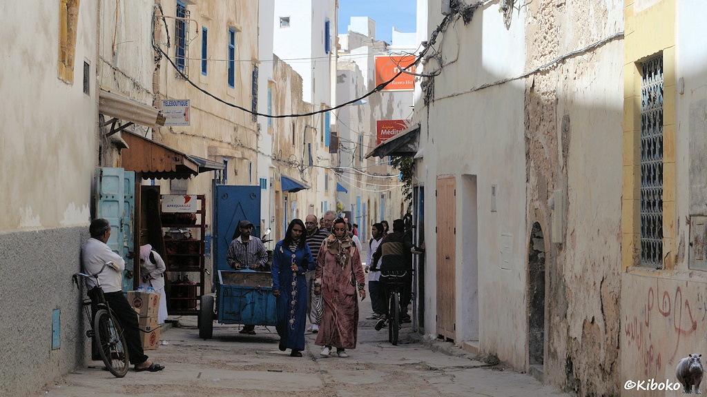 Das Bild zeigt eine Gasse zwischen zwei- und dreistöckigen weißen Häusern mit blauen Türen und Fenstern. Auf der Gasse kommen zwei Frauen im blauen und rosa Mantel entgegen. Dahiner kommt ein Mann mit blauem Handwagen und weitere Männer. Am linken Bildrand lehnt ein Mann mit seinem Fahrrad an der Hauswand. Rechts bröckelt der Putz von der Wand.
