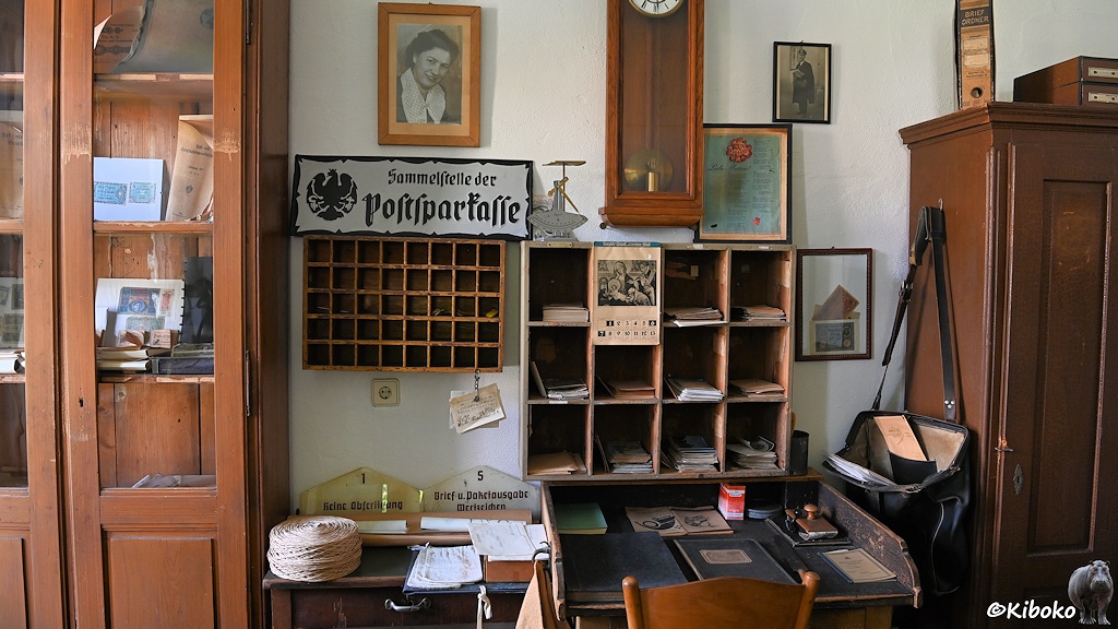 Das Bild zeigt die Innenansicht einer Poststelle. An der Wand sind hölzerne Sortierfächer. Davor steht ein hölzernes Pult. An einem Schrank hängt eine offene Tasche des Postzustellers. An der Wand hängt ein Schild mit der altzdeutschen Schrift: Sammelstelle der Postsparkasse.