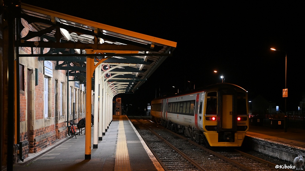 Das Bild zeigt einen zweiteiligen, weißen Dieseltriebwagen mit roten Türen und gelber Front nachts in einem zweigleisigen Bahnhof. Am linken Bildrand ist das Empfangsgebäude mit der Bahnsteigüberdachung. Der Triebwagen steht auf dem zweiten Gleis und zeigt die Rücklichter.