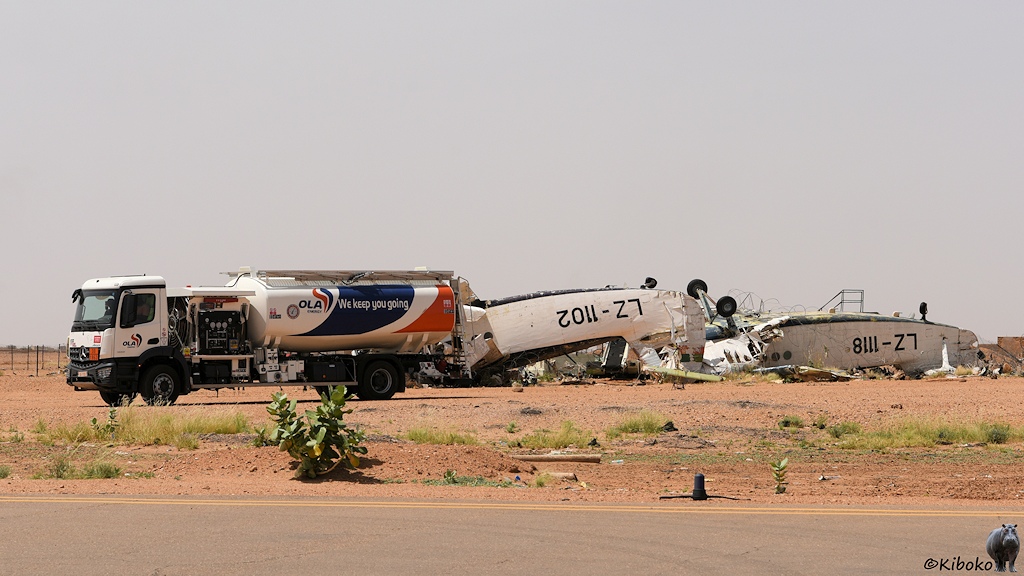 Das Bild zeigt einen modernen weißen Tankwagen der Firma OLA mit weiß-blau-orangem Tank. Auf dem Tank steht: We keep you going. Dahinter liegen zwei alte Flugzeuge auf dem Rücken mit den Anschriften LZ-1102 und LZ-1118.