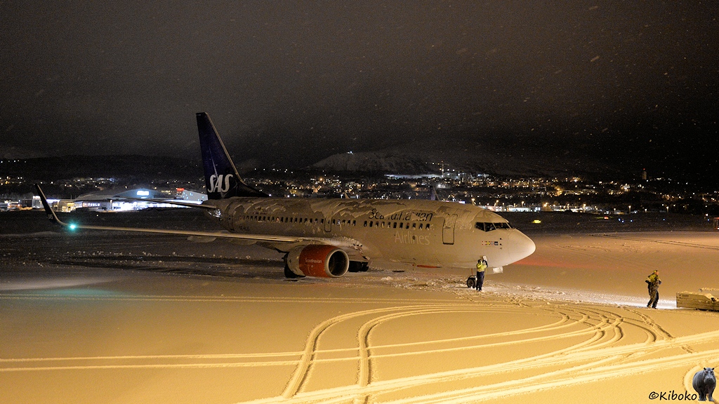 Das Bild zeigt einen kleinen Düsenflieger mit blauem Leitwerk und roter Triebwerksverkleidung auf einem verschneiten Flugfeld. Schnell liegt auf dem Rumpf und Tragflächen des Flugzeugs. Auf dem Flugfeld sind Fahrzeugspuren. Im Hintergrund sind die beleuchteten Häuser einer Stadt. Am oberen Bildrand belegen unscharfe helle Streifen dichten Schneefall.