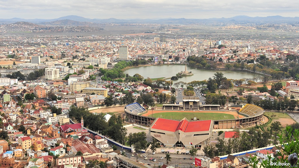 Das Bild zeigt eine Stadt von einem Hügel. Unten rechts ist ein Fussballstadion mit einem Eingangsportal mit rotem Dach. Die Tribüne zieht sich auf beiden Seiten herum. Auf der Rückseite ist dann ein kleinere Triüne. Dahinter führt eine Straße auf einen See mit Insel und dem Obelisk mit Goldengel. Die Häuser breiten sich ein einer Ebene aus, die durch Berge in großer Entfernung begrenzt ist.
