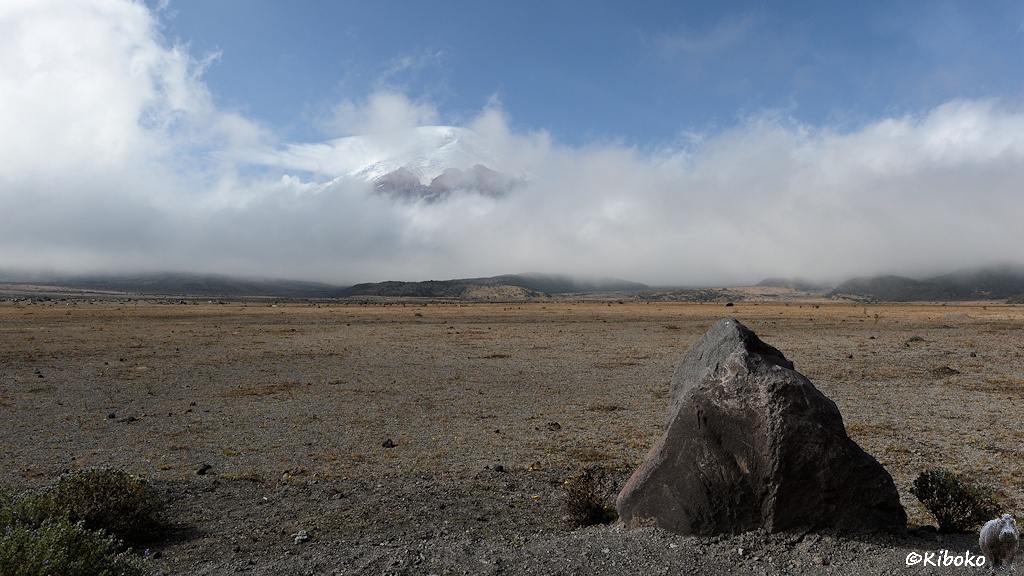 Das Bild zeigt einen dreckigen Stein mit der Spitze nach oben in einer weiten Schotterebene. Im Hintergrund ist ein Vulkan mit Schneekuppe der über die eine umgebene Wolkendecke herausschaut.