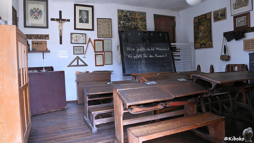 Das Bild zeigt die Innenansicht einer Schule mit vier hölzernen Schulbänken in unterschiedlichen Höhen. Vor der Wand steht eine Tafel mit der Kreideaufschrift: Wie gefällt Dir das Museum. Am der Wand hängen Tabellen, Bilder und ein Kruzefix.