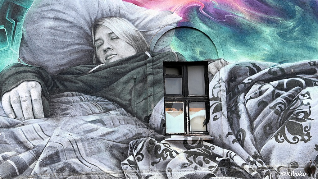 Das Bild zeigt ein Wandgemälde einer schlafenden Frau mit Kissen und gemusterten Decke in grau-weiß. Im Hintergrund sind grüne und violette Verläufe. Im Zentrum wird da sBild durch ein Fenster mit schwarzem Rahmen unterbrochen.