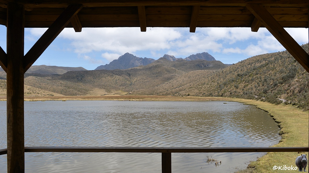 Das Bild zeigt den Blick aus einer überdachten Plattform am rechten Ufer auf einem See. Im Hintergrund ist ein fesliges Bergmassiv. Im Vordergrund wird das Bild eingerahmt durch einen Stützbalken, ein Stück vom Dach und dem Geläner.