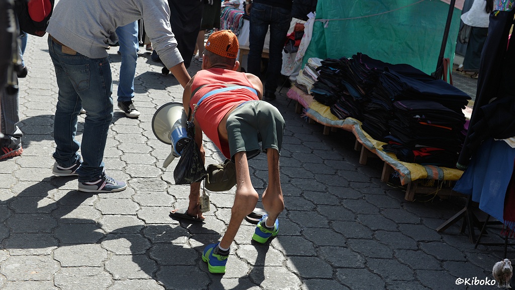 Das Bild zeigt einen Mann auf allen Vieren auf einen gepflasterten Weg zwischen Marktständen. Die Beine und Becken sind sehr dünn. Die Kniegelenke sind defomiert. Die Hände stecken in Latchen. An einem Gürtel trägt er ein Megaphon, dass neben dem linken Arm baumelt. Ein Mann bückt sich und spndet etwas.