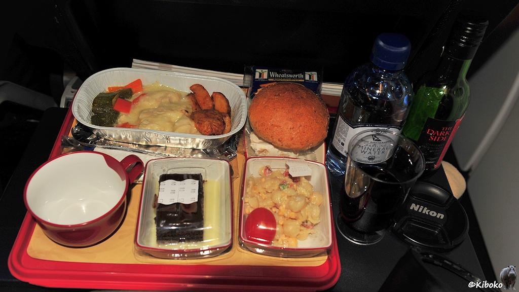 Das Bild zeigt ein rotes Tablett auf einem runtergklappten Tisch im Flugzeug. Auf dem Tablett stehen ein offene Alushale mit ein paar Bratkartoffeln, Gurken, Karotten und etwas undefinierrbaren gelben Brocken. Des weiteren sind dort ein trockenes Brötchen, eine leere Tasse, ein Stück Kuchen und eine Schalte mit gelbem Nudelsalat und Tomate. Neben dem Tablett steht eine Wasser- und eine Weinflasche mit der Aufschrift: Dark Side. Des weiteren liegt ein runder schwarzer Objektivdeckel daneben.