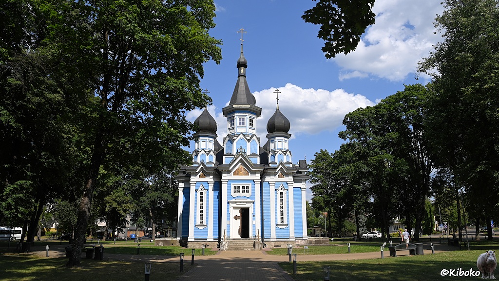 Das Bild zeigt eine blaue Holzkirche mit weißen Säulen. Das Dach und die drei Türmchen sind dunkelgrau eingedeckt. Dir Kirche steht in einem Park zwischen hohen Bäumen.