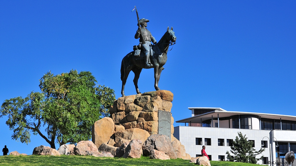 Ein Denkmal eines bewaffneten Reiters auf einem Pferd steht auf einer Wiese vor einem flachen Gebäude.