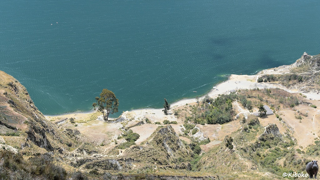 Das Bild zeigt einen Ausschnitt vom vorderen Rand des türkisfarbenen Sees. Ein heller schmaler Streifen am See ist ein Strand. Ganz klein wind ein Kajak und ein paar Menschen am Strand sichtbar. Am linken oberen Bildrand sind zwei Kajaks auf dem See.