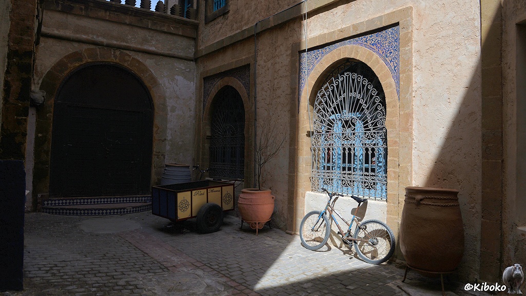 Das Bild zeigt einen Innenhof im Schatten. Auf der rechten Seite scheint in ein dreieckiges Feld. Darin steht ein rostiges Fahrrad ohne Lampen und ohne Schutzbleche. Darüber wird von der Sonne ein blaues Fenster mit Rundbogen und feinem Schmiedegitter angestrahlt. Über dem Rudnbogen sind feine blaue Muster an der Fassade. Links sind im Schatten ein weiteres Fenster und ein breites, braunes Tor. Im Innenhof steht noch ein Handwagen mit Gummireifen und ein Bäumchen in einem Tongefäß.