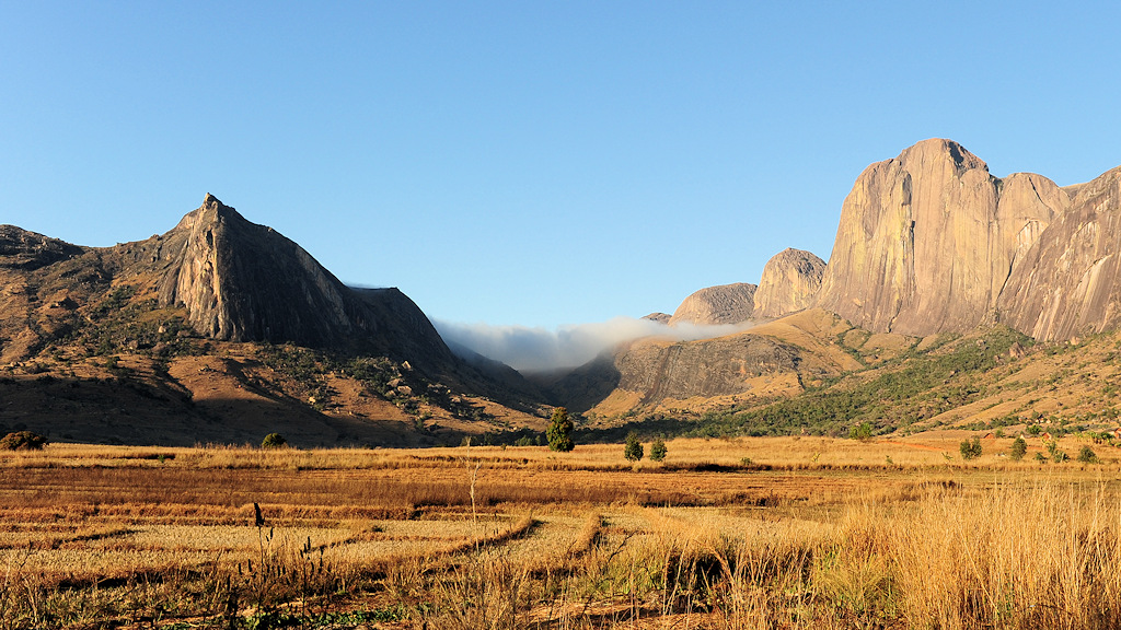Das Bild zeigt sonnenbeschienenes Tal. Links ist ein Berg mit einer spitzen Felsspitze im Schatten. Links ist Bergmassiv mit hochen Felswänden. Dazwischen hängen Wolken an einer Paßhöhe. Im Talboden sind trockene, abgerntete Reisfelder.