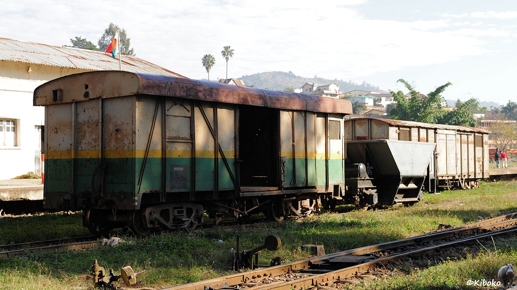 Das Bild zeigt vierachsigen, gedeckten Güterwagen mit wulstigen Dach. Der Güterwagen ist oben beige, unten dunkelgrün mit trennendem gelben Streifen. Dahinter steht ein tricherförmiger wagen und auf dem nächsten Gleis ein grauer, vierachisger gedeckter Güterwagen.