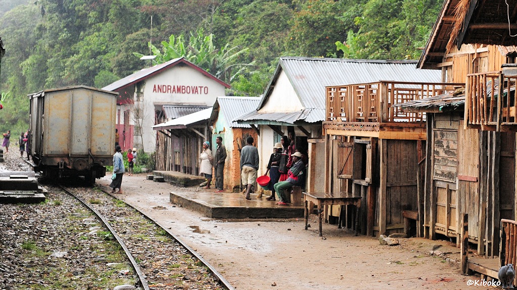 Das Bild zeigt einen kleinen Bahnhof. Auf dem Nebengleis steht ein grauer, gedeckter Güterwagen. Neben dem Gleis stehen Holz- und Steinhäuser mit Wellblechdächern. Im Hintergrund ist ein weißes, einstöckiges Gebäude mit dem Bahnhofsnamen.