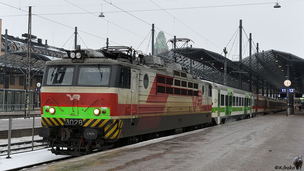 Eine rot-weiße Elektrolomkomotive mit güner Kontrastfläche steht vor einem Zug aus grün-weißen und rot-weißen Wagen vor einer Bahnhofshalle.