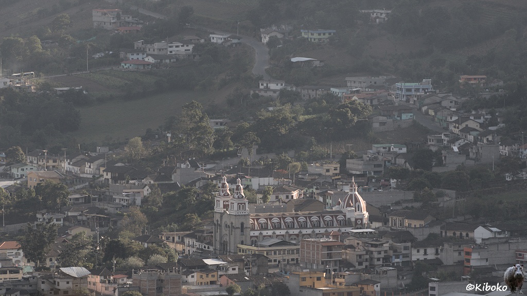 Das Bild zeigt einen Ort im Gegenlicht. Im Zentrum steht eine große Kirche mit zwei Türmen am Eingangsportal und hinten eine große Kuppel.