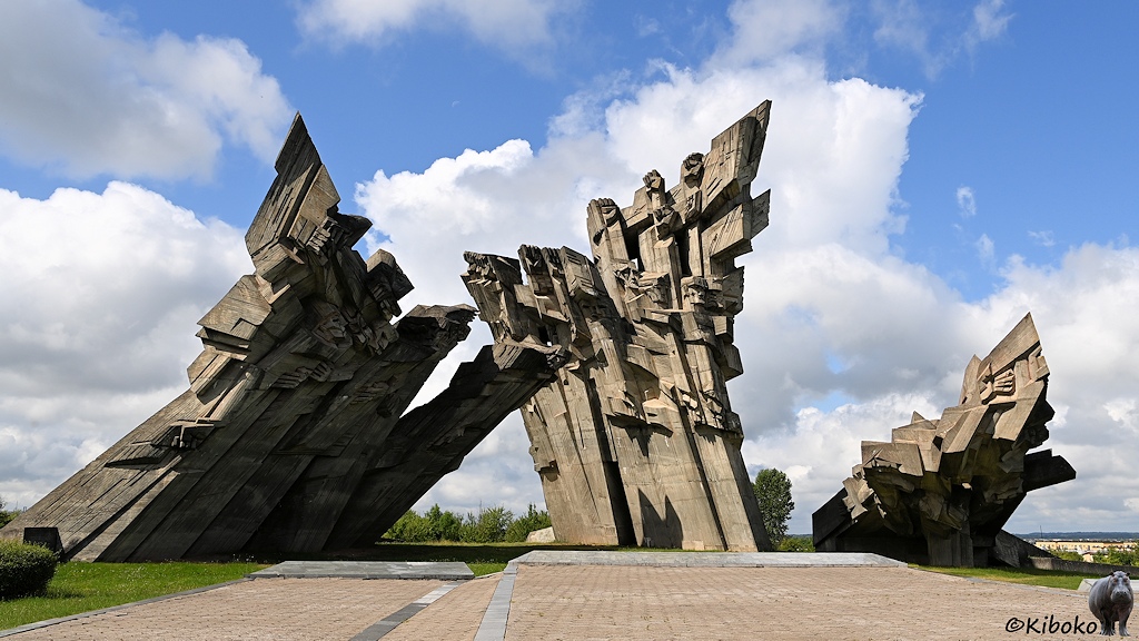 Das Bild zeigt ein Mahnmahl aus drei geneigten Betonklötzen aus jweils 5 aneinandergereihten Stelen. An den Spitzen der Stelen sind Figuren und Köpfe. Der Linke Betonklotz ist nach rechts geneigt, der mittlere nach links und der rechte ist sehr stark nach vorn geneigt. Der Vordregrund ist gepflastert.