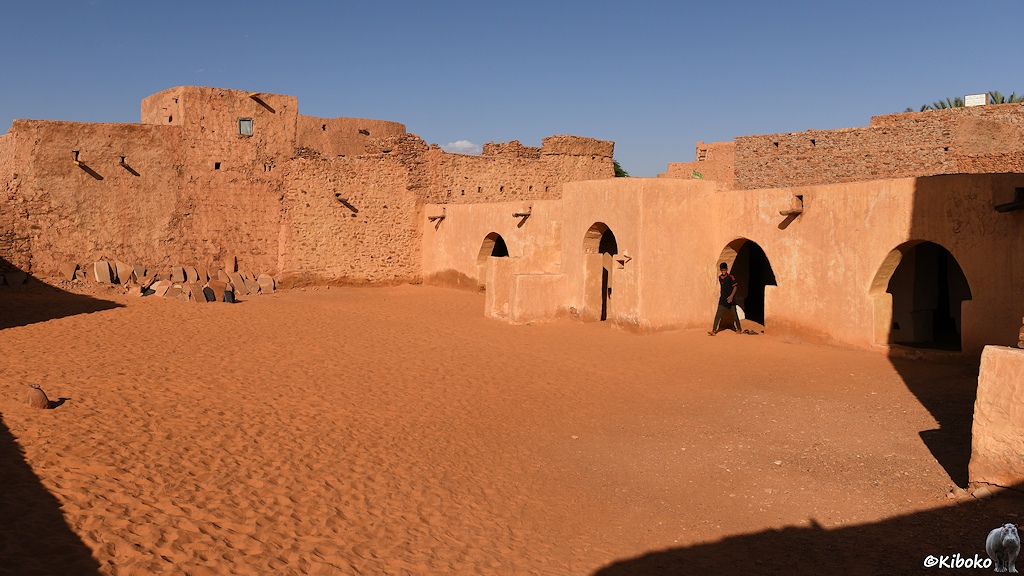 Das Bild zeigt den sandigen Innenhof einer Moschee. Auf der rechten Seite ist ein einstöckiges Gebäude mit vier pilzförmigen Eingängen. Dahinter ist eine Wand aus rötlichem Natursteinen.