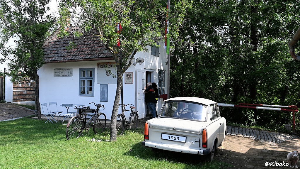 Das Bild zeigt ein kleines weißes Gebäude mit einem rot-weißen Schlagbaum davor. Vor dem Schlagbaum steht ein grauer Trabant mit dem Kennzeichen 1989.