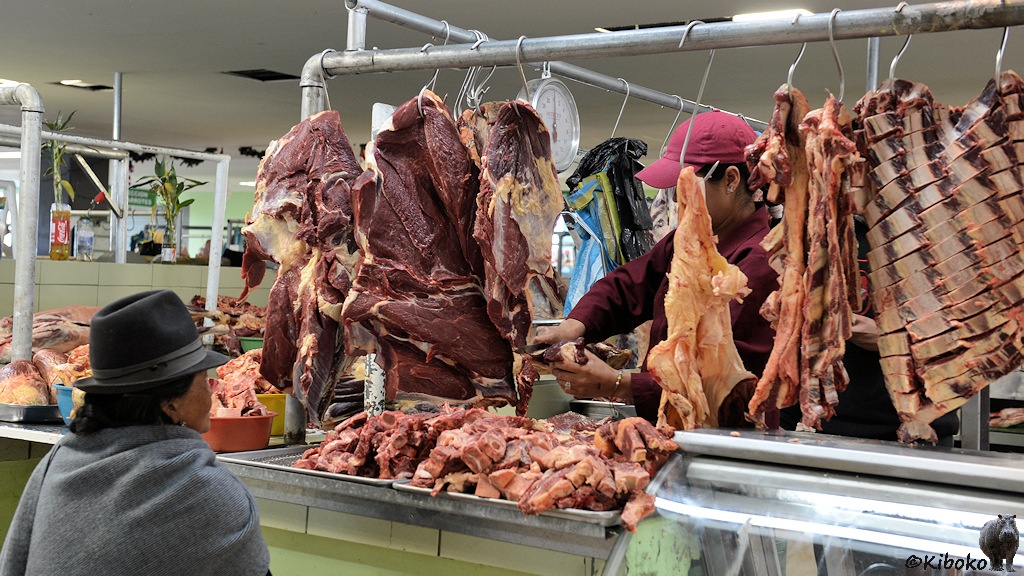 Das Bild zeigt die eine Frau im grauen Poncho mit grauem Hut von hinten. Sie schaut zu, wie ein Schlachter mit rotem Hemd und roter Mütze ein Stück fleisch von einem großen Brocken abschneidet. Daneben hängen weitere Fleischbrocken. Kleinere Stücken liegen in silbernen Schüsseln auf der Tresen.