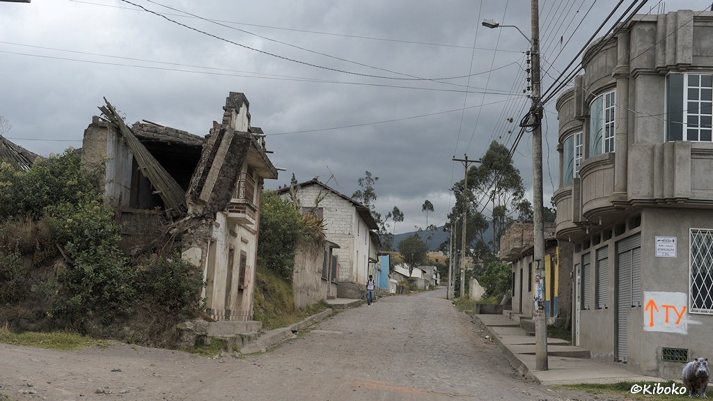 Das Bild zeigt eine schmaler Kopfsteinpflasterstraße. An der linken Seite steht eine Hausruine mit eingestürzten Gibel und Dach. Am rechten Bildrand steht ein modernes Haus aus Beton mit vergitterten Fenstern und heruntergelassenen Jalousien.