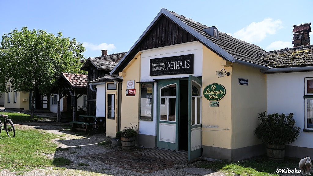 Das Bild zeigt eine Reihe kleiner Einstöckiger Steingebäude im Freilichtmuseum. Im Vordergrund ist ein beigefarbenes Gebäude mit einer offenen Flügeltür auf der Giebelseite. Darüber steht auf einem Schild: Gemischtwarenhandlung und Gasthaus. Rechts neben der Tür ist ovales grünes Emailleschild mit der Aufschrift: Liesinger Bier.